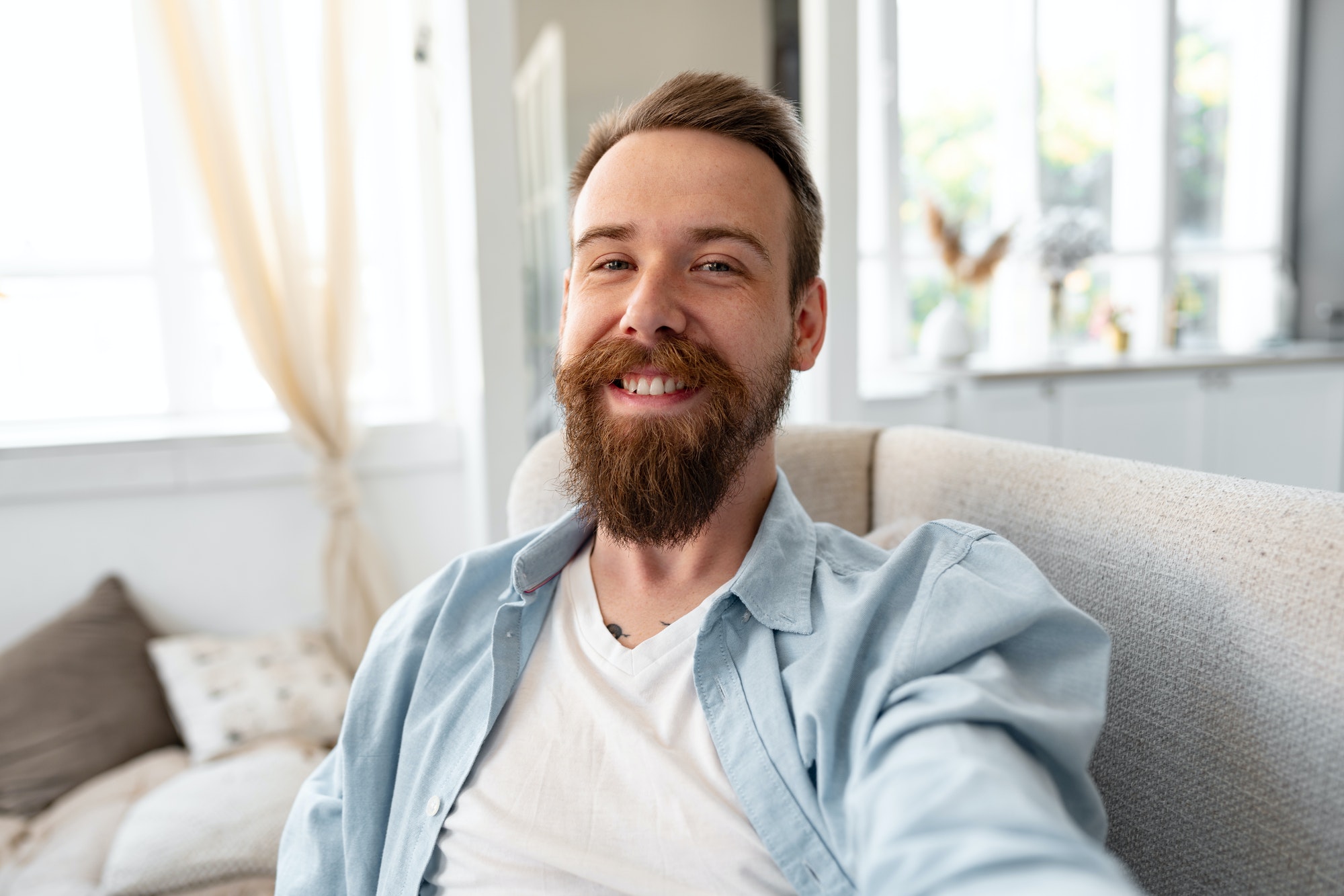 Bearded happy young man smiling and taking selfie photo while sitting on sofa at home.